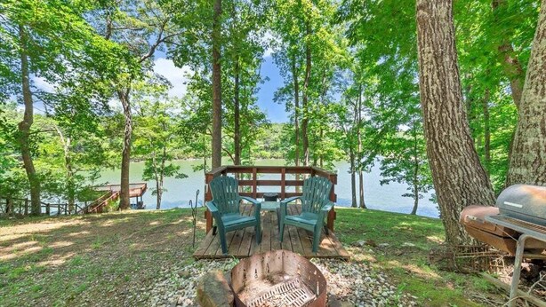 Un terrain de camping équipé d’un foyer, de deux chaises vertes, d’un barbecue et d’une terrasse en bois donnant sur un lac.