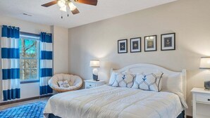 Une chambre avec un grand lit, un ventilateur de plafond, une fenêtre avec des rideaux bleus, une chaise, une table de chevet avec une lampe et des tableaux encadrés accrochés au mur.