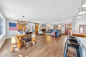 Amplia sala de estar con mesa de comedor, sillas de madera, sofá y una cocina con mostrador de madera y taburetes de bar.