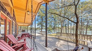 A wooden deck with red Adirondack chairs, a hammock, and a view of a lake and trees. - Lake Harmony - Lakefront Home w/ Private Dock, & Fire Pit (Huddleston)