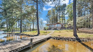 A cabin with a dock extending into a clear stream, surrounded by tall trees and a grassy area. - Lake Harmony - Lakefront Home w/ Private Dock, & Fire Pit (Huddleston)