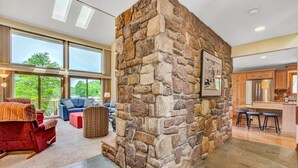 A spacious living area with a stone accent wall, large windows offering a view of greenery, and a modern kitchen with bar stools. - Dancing Water - Sleeps 10, Private Dock, Game Room & Hot Tub (Moneta)