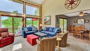 A spacious living room with a large window overlooking a lake, a dining area with wooden furniture, and a red and blue sofa set. - Dancing Water - Sleeps 10, Private Dock, Game Room & Hot Tub (Moneta)