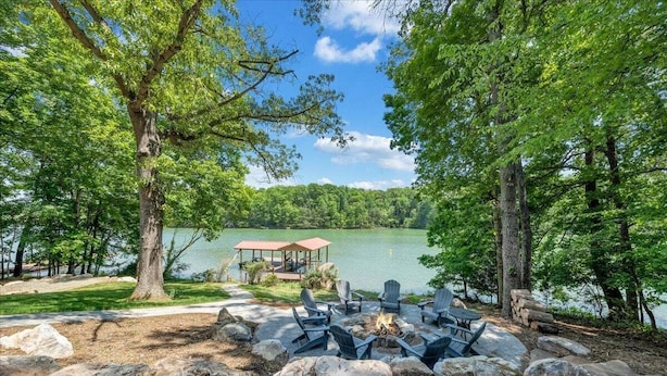 Ein Sitzbereich am See mit Feuerstelle und Adirondack-Stühlen, umgeben von Bäumen und einem Steg.