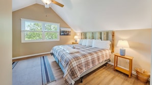 Ein Schlafzimmer mit schrägen Wänden, einem Bett mit kariertem Überwurf, einem Deckenventilator, einem Fenster mit Jalousien, einem Nachttisch mit Lampe und einem Holzboden.