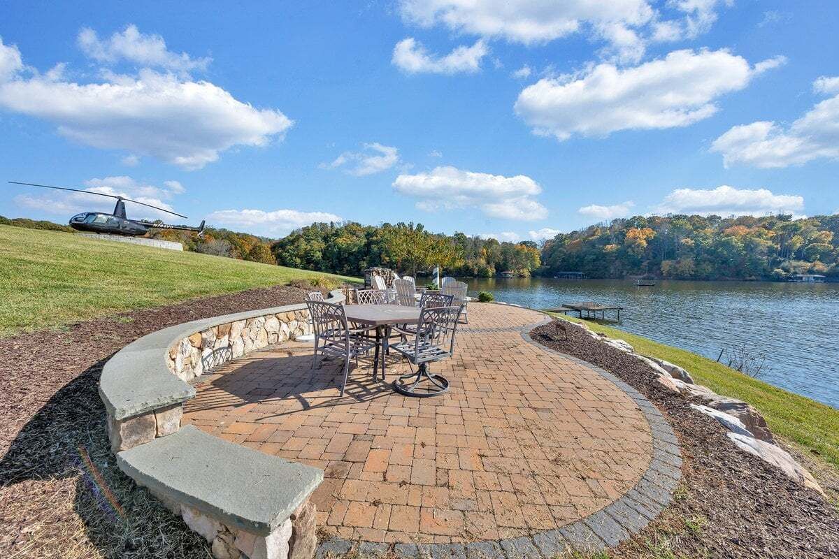 A patio with a circular brick design, outdoor furniture, and a view of a lake with a helicopter in the background.