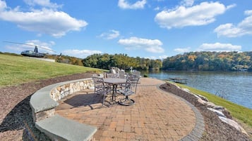A patio with a circular brick design, outdoor furniture, and a view of a lake with a helicopter in the background.