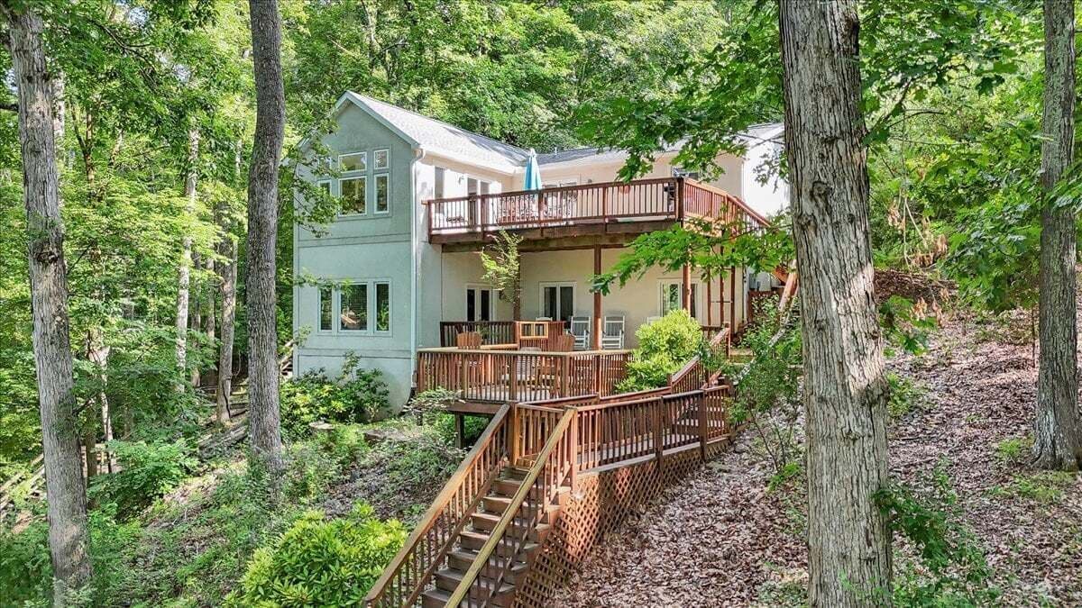 A two-story cabin with a wooden deck and stairs, surrounded by dense forest.