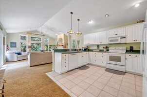 Una cocina amplia con gabinetes blancos, microondas y estufa. Hay un comedor con sofá y alfombra.