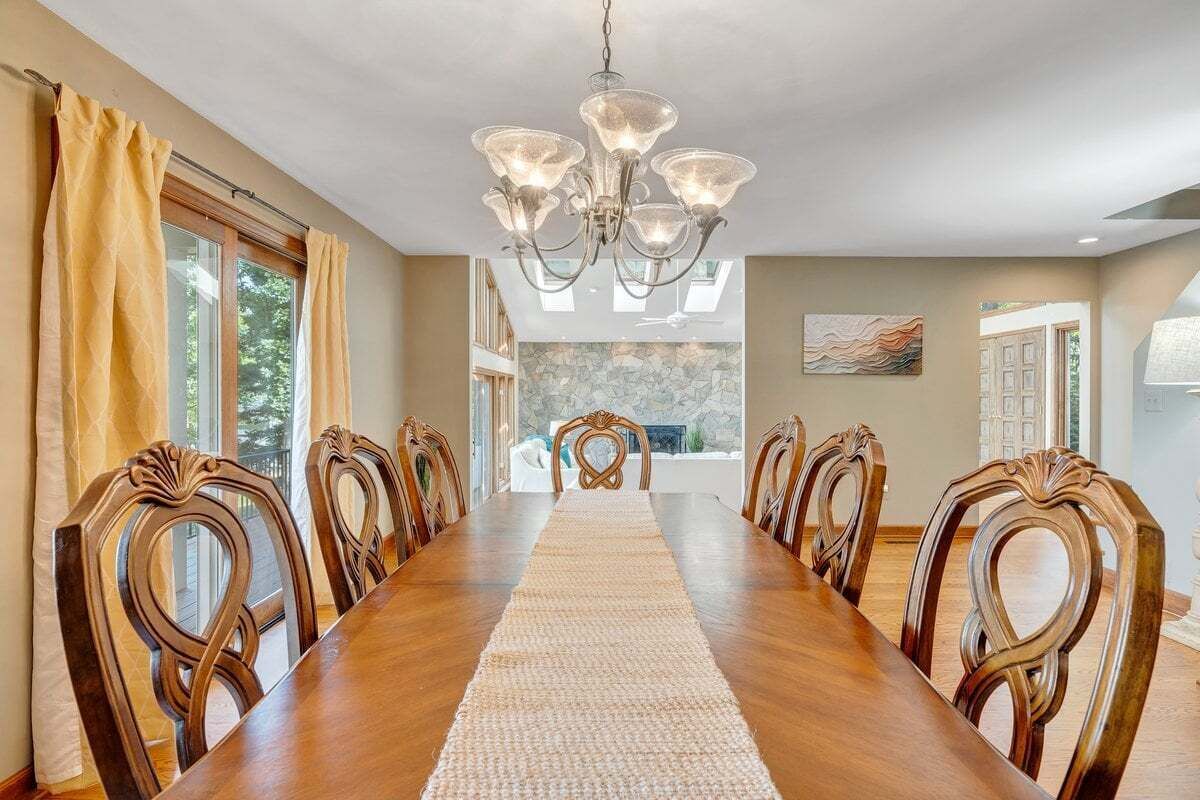 A dining room with a long wooden table, ornate chairs, a chandelier, and a view of a stone wall and trees through large windows.
