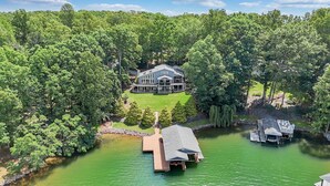 Une grande maison entourée d’une forêt dense, un quai et un petit abri pour bateaux au bord d’un lac verdoyant.