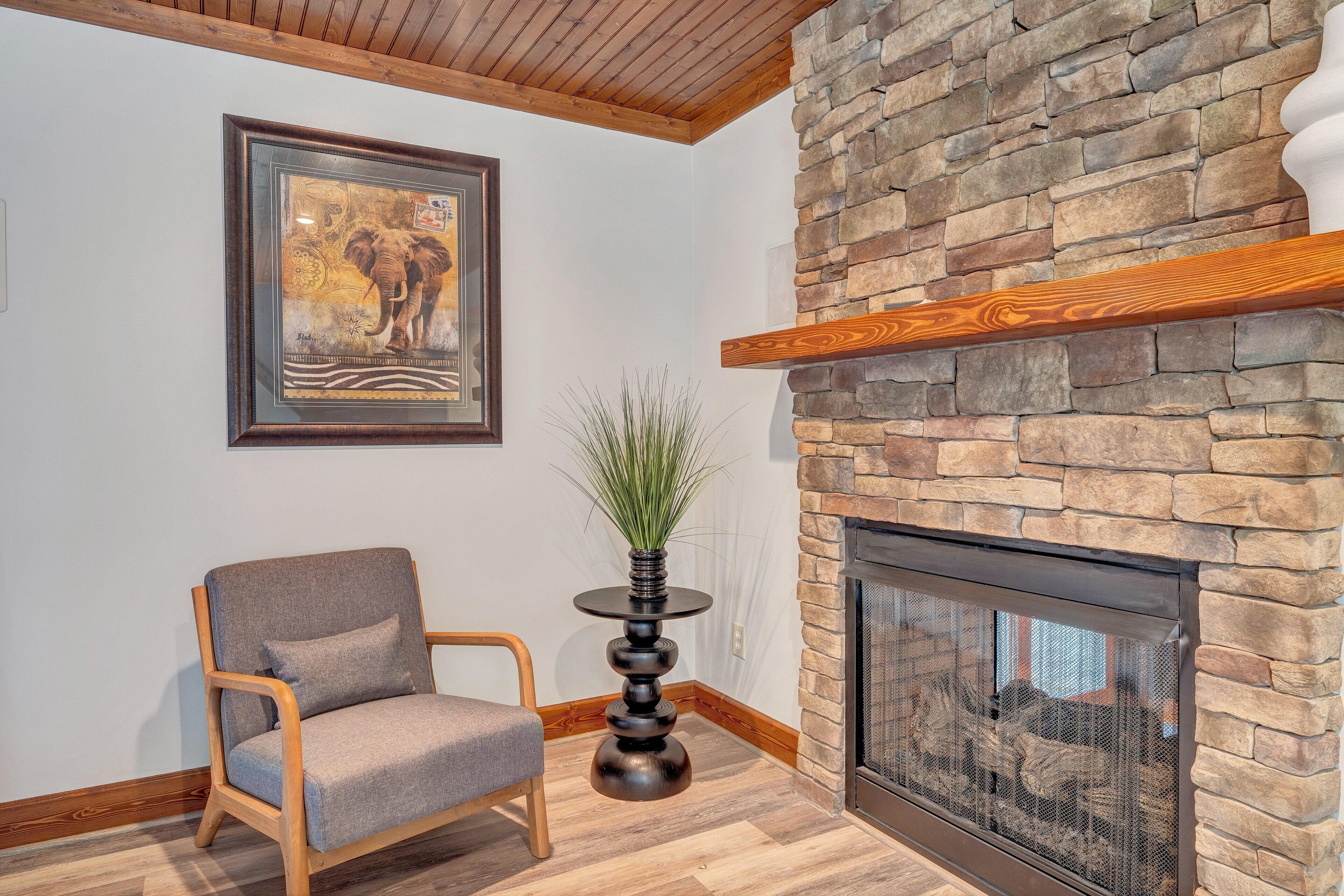 A room with a stone fireplace, a framed picture on the wall, a chair, and a small table with a plant.