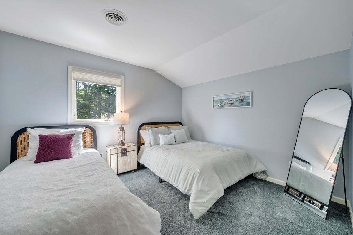 Een slaapkamer met twee bedden, een raam, een nachtkastje, een lamp, een spiegel en een lijst met een afbeelding aan de muur.