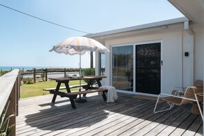 A wooden deck with a picnic table and umbrella, a glass door leading to a covered area, and a view of the sea. - The Nook Studio ~ 1 bedroom ~ stunning ocean views (Aldinga Beach)