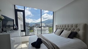 A modern hotel room with a large bed, a desk, and a view of a cityscape through large windows. - Luxury Studio in Harmony - Best Area Quito, Pool, Gym & Fast WiFi (Quito)