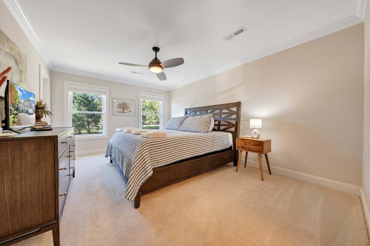 A bedroom with a bed, a dresser, a nightstand, a ceiling fan, and windows.