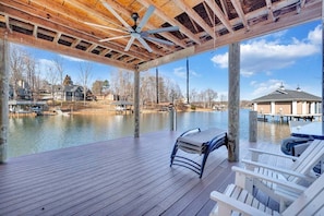 A wooden deck with a ceiling fan, overlooking a calm body of water with docked boats and houses in the background. - Serenity Shores - Private Lakefront Home With Dock (Moneta)