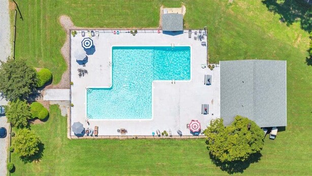 Eine Vogelperspektive auf einen rechteckigen Pool mit einer umlaufenden Terrasse, umgeben von Bäumen und einem Gebäude.