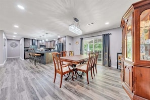 A spacious dining area with a wooden table and chairs, a kitchen in the background, and a large wooden cabinet with glass doors. - Lakeview Village East - Community Pool, Tennis & Boat Ramp! (Moneta)