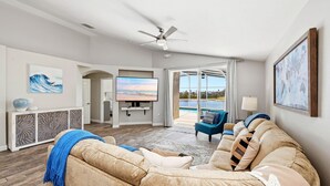 A living room with a beige sofa, a television, and a view of a pool through glass doors. - Golden Pond Lakehouse. Tranquil lake retreat with charm, designer style & private pool by Disney (Kissimmee)