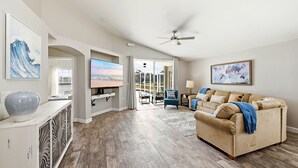 A spacious living room with a large flat-screen TV, a beige sofa, and a ceiling fan. - Golden Pond Lakehouse. Tranquil lake retreat with charm, designer style & private pool by Disney (Kissimmee)