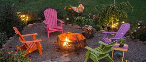A fire pit surrounded by colorful Adirondack chairs, a small table, and potted plants.