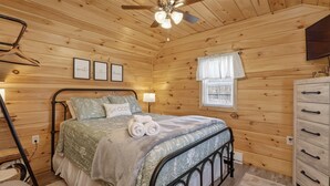 Une chambre à coucher dans une cabane en bois, avec un lit, une commode, un ventilateur de plafond et une fenêtre avec des rideaux.