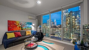 A modern living room with a city view, a sofa with colorful cushions, a coffee table, and a large window. - Convenient CBD Retreat in the Best Location! 2 Bed | 2 Bath | 2 Parking (Melbourne)