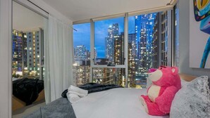 A hotel room with a large window offering a city view, a bed with a teddy bear, and a cityscape at dusk. - Convenient CBD Retreat in the Best Location! 2 Bed | 2 Bath | 2 Parking (Melbourne)
