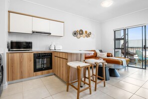 A modern kitchen with a microwave, oven, and coffee maker, a bar stool area, and a sofa with a view to the outside. - Ballito Hills 887 - One Bedroom Apartment, Sleeps 2 (Durban)