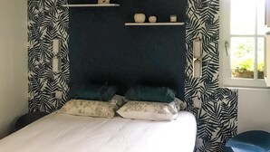 Une chambre à coucher comprenant un lit, des oreillers, une table de chevet, une étagère avec des objets décoratifs et une fenêtre donnant sur un paysage verdoyant.