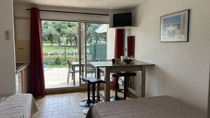 A room with a bed, a desk with a chair, a TV mounted on the wall, and a sliding glass door leading to an outdoor area. - Pleasant studio cabin apartment in residence with pool near Richelieu beach, Cap d'Agde (LE CAP D AGDE)