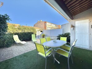 Une terrasse avec une table ronde et quatre chaises, entourée de gazon synthétique et d’un chemin de gravier.