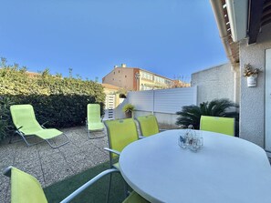 Une terrasse extérieure avec une table ronde et six chaises vertes, entourée d’une haie et d’un bâtiment en arrière-plan.
