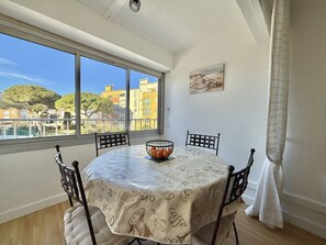 A dining area with a round table, four chairs, a bowl of oranges, and a view of buildings through large windows. - CAP D'AGDE - Les Falaises // Réf 0144 (CAP D'AGDE)