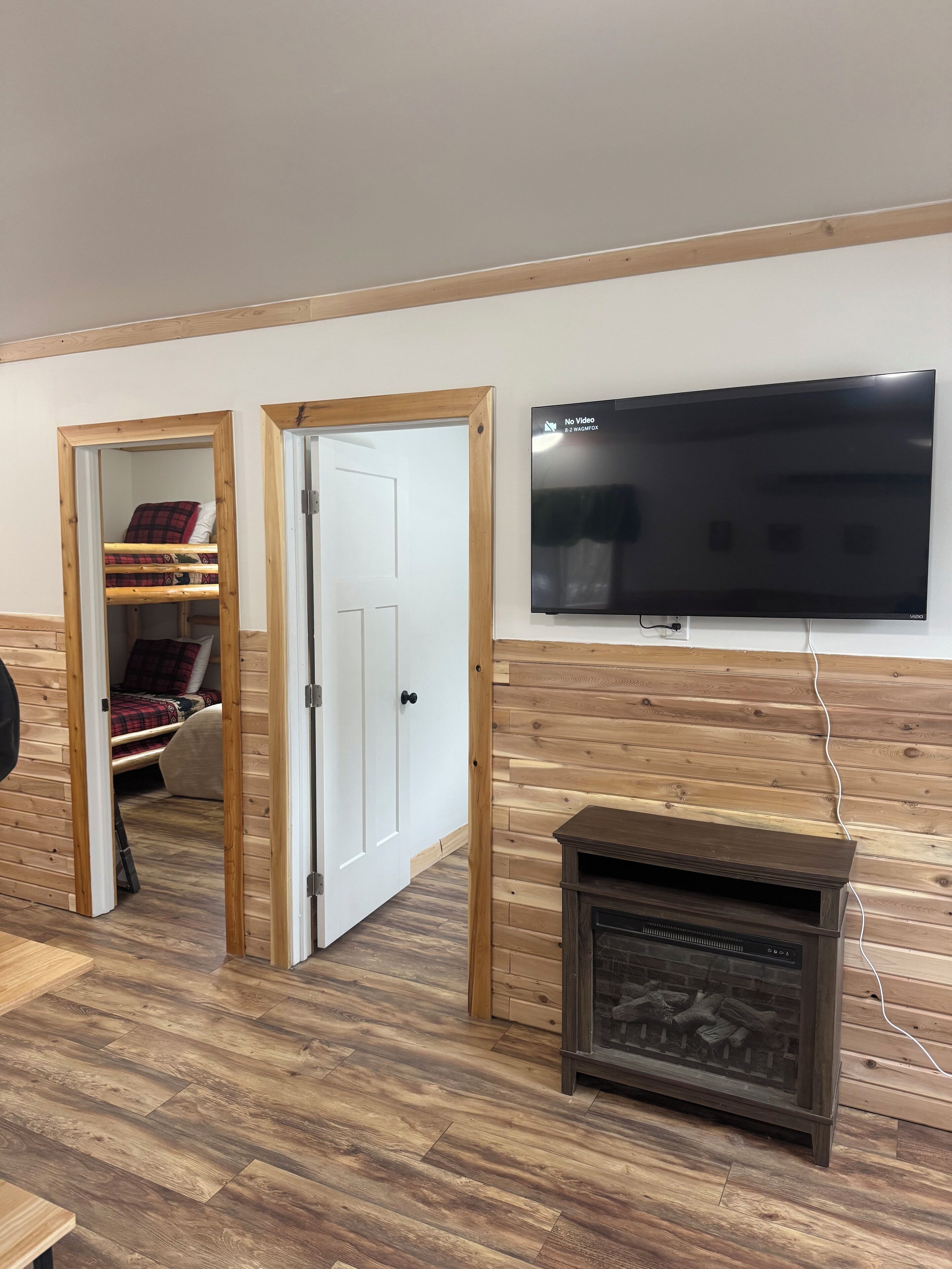Una habitación con piso de madera, televisor montado en la pared, chimenea y puerta que da a una cama con colcha a cuadros roja y negra.