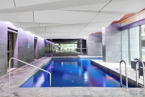 A modern indoor swimming pool with clear glass walls, a tiled floor, and a metal pool ladder. - Stylish Melbourne CBD Getaway - Pool & Gym Access (Melbourne)