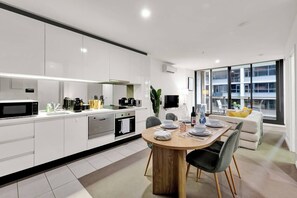 A modern kitchen with white cabinetry, a dining area with a wooden table, and a large window offering a view of the city. - Stylish Melbourne CBD Getaway - Pool & Gym Access (Melbourne)