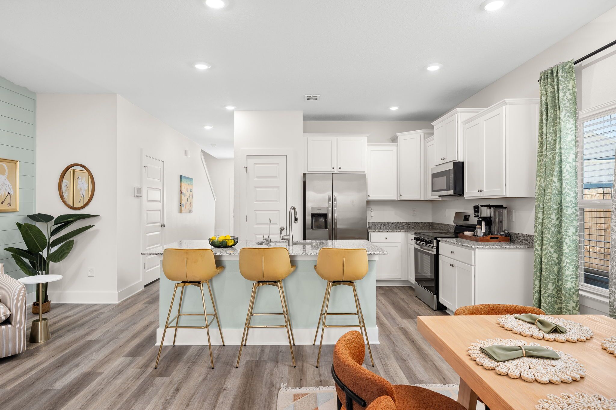 Una cocina moderna con armarios blancos, un refrigerador de acero inoxidable y un microondas. Hay tres taburetes de bar con patas doradas y una mesa de comedor de madera con individuales.