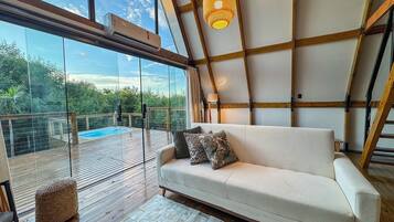 Interior moderno com sofá, luminária pendente e parede de vidro com vista para uma piscina.
