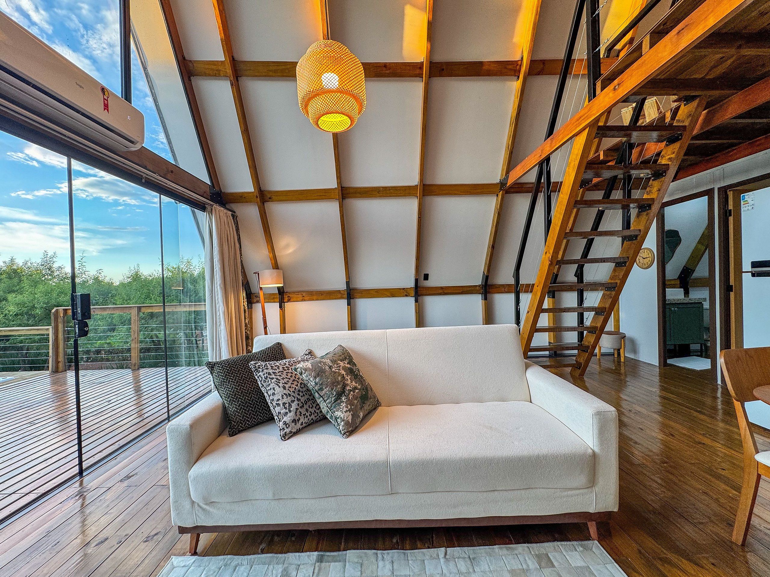 Un intérieur moderne avec un plafond en bois, un canapé blanc agrémenté de coussins à motifs et une balustrade en verre donnant sur une terrasse.