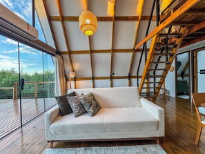 Un interior moderno con techo de madera, un sofá blanco con cojines estampados y una barandilla de vidrio que da a una terraza.