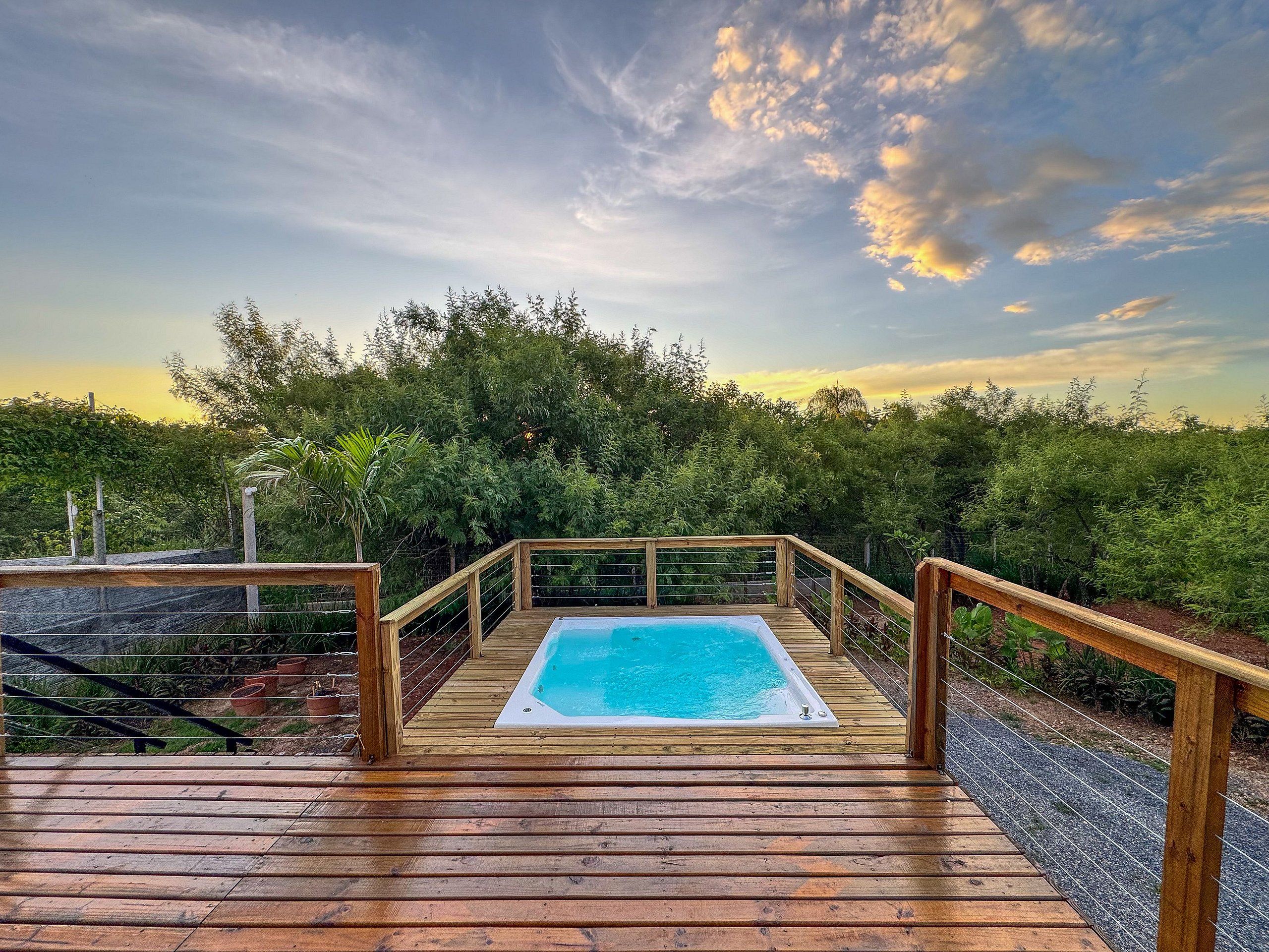 A wooden deck with a hot tub, surrounded by greenery and a clear sky.