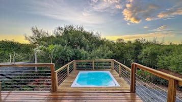 A wooden deck with a hot tub, surrounded by greenery and a clear sky.