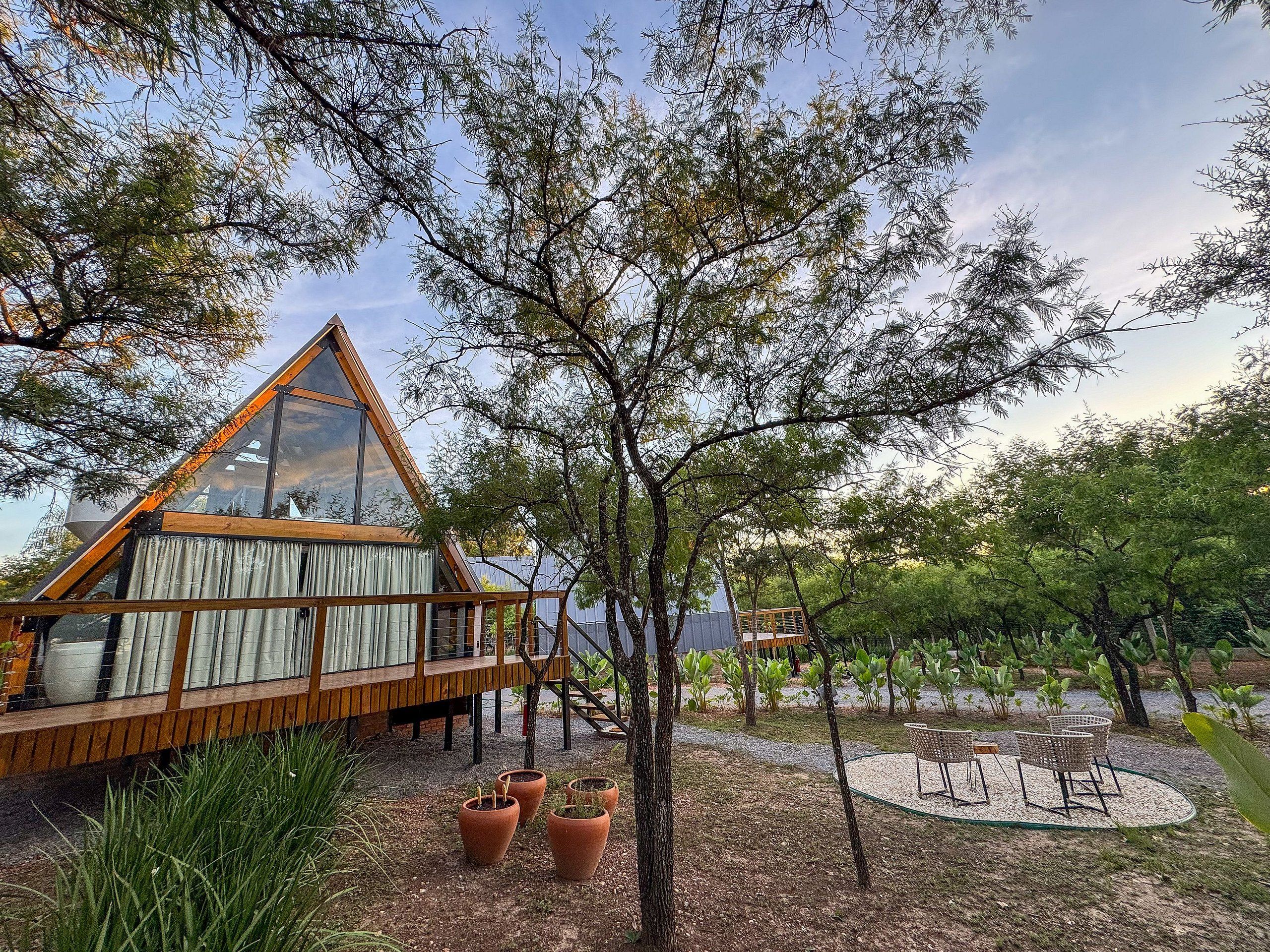 Uma cabana em A moderna com varanda, rodeada por árvores e uma área de cascalho com plantas em vasos e móveis de exterior.