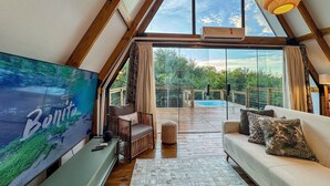 A spacious loft bedroom with a large window, a cozy seating area, and a view of a swimming pool. - Chalé Folk Village Moderno com jacuzzi (Bonito)