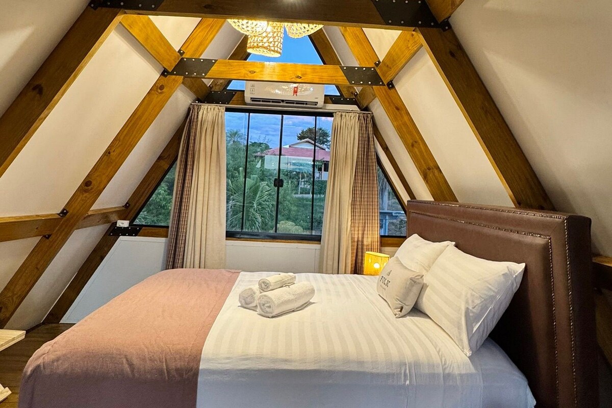 Ein Schlafzimmer mit schrägen Wänden, einem Bett mit Kissen und Decke, einem Nachttisch mit Lampe sowie einem Fenster mit Vorhängen.