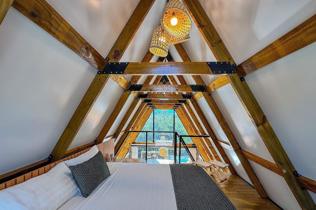 Quarto de sótão com cama, duas almofadas, luminária pendente e vista para o exterior.