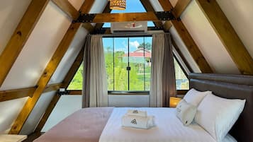 A bedroom with a sloped ceiling, a bed with white linens, a window with curtains, and a view of greenery outside.