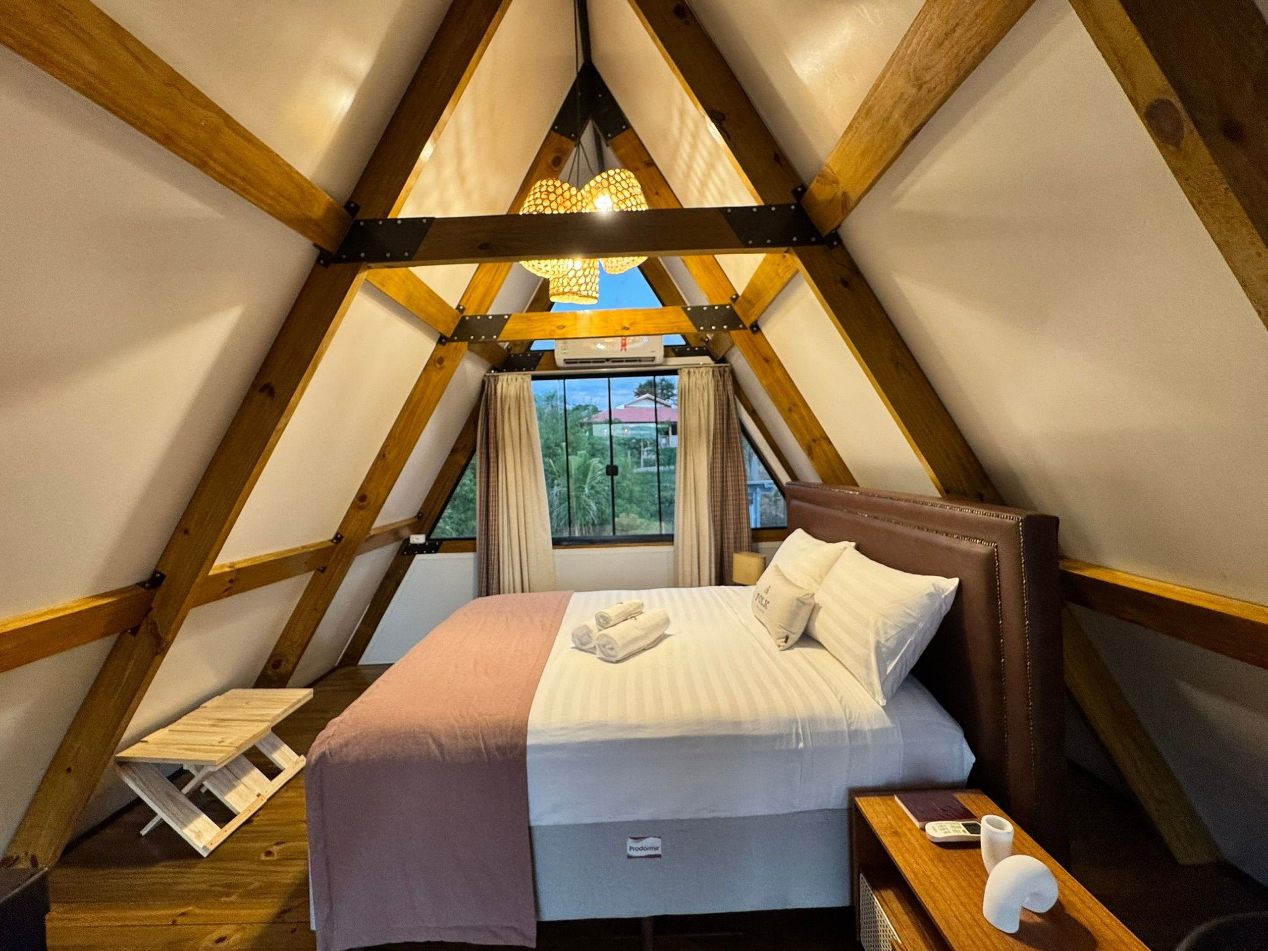 Une pièce triangulaire comprenant un lit, des poutres en bois, une fenêtre avec des rideaux et un luminaire suspendu.